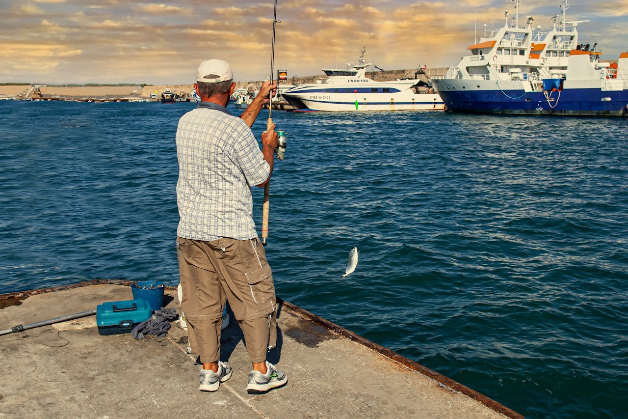 A fisherman reels in a catch by the pier at LAmetlla de Mar, Spain with boats in the background.